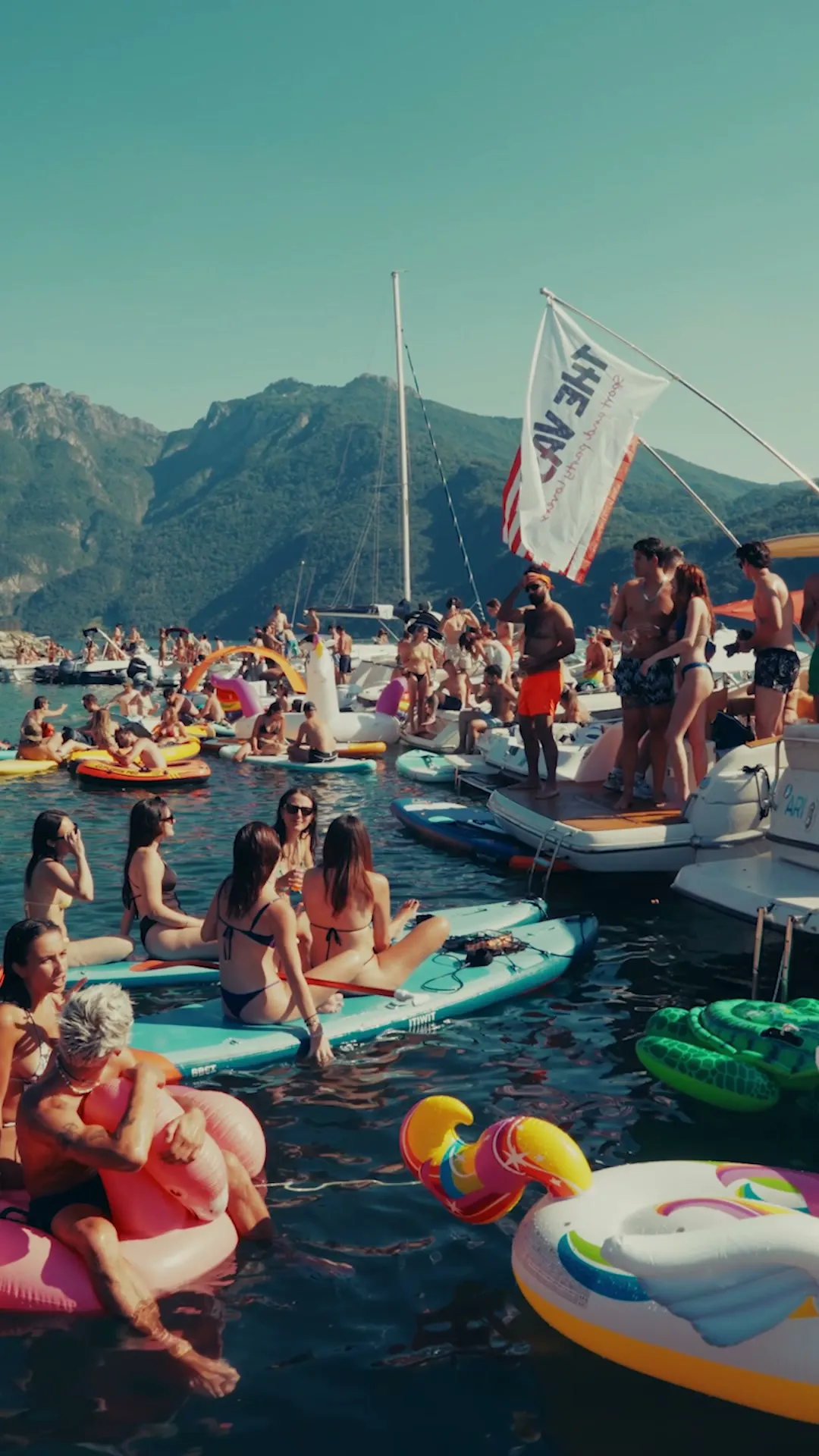 Folla in acqua su gonfiabili e tavole da SUP, con le barche e le montagne sullo sfondo